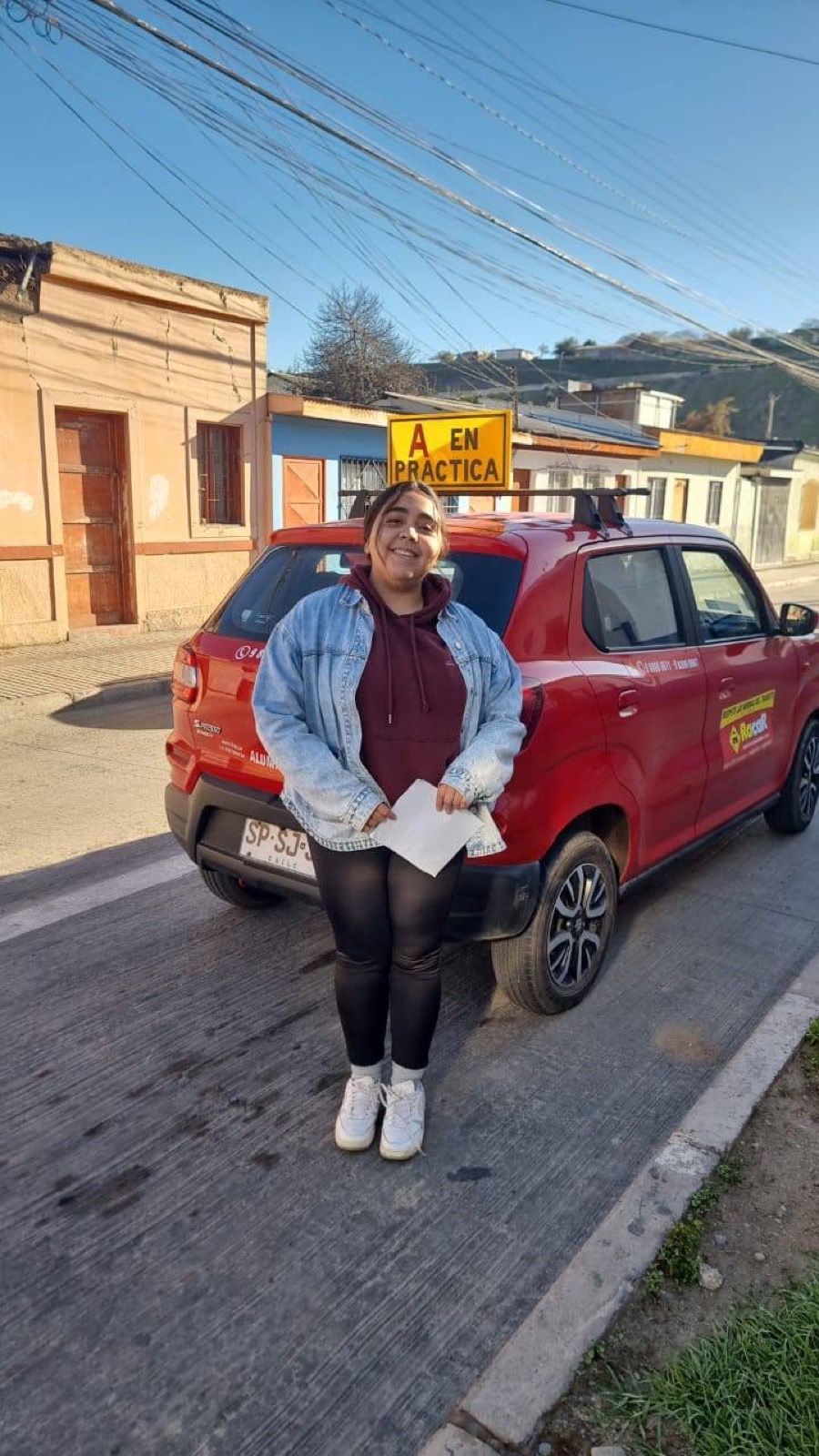 Alumna sonriendo junto al auto rojo de la escuela con el cartel A en práctica, en Vallenar